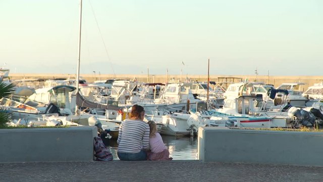 Little Girl with her Mother Reading a Touristic Map while Sitting on the Steps near Yachts and Boat Parking in Harbor at Sunset. They Travelling around Ischia Island in Italy.