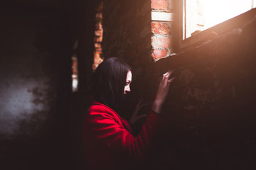 stylish girl with black hair in a red coat and long skirt looks out the window in an old abandoned building