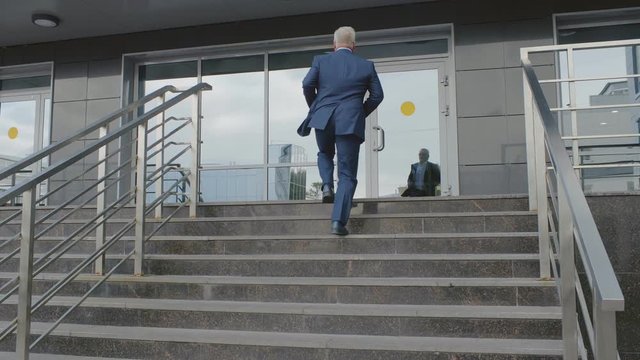 Confident senior caucasian businessman in suit and tie going up the stairs of a business centre and enter glass doors of office center. Office worker go to work place in the morning