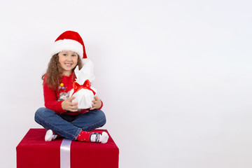 A little girl in a Santa Claus hat sitting on a red cube in the Studio with a gift in her hands, on a white background. The concept of a happy childhood. Blank space for text. The layout for the cards