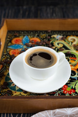 A white cup of coffe staying on a fancy tray with traditional decorative painting style - Ukrainian Petrykivka - on dark wooden background
