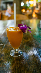 A healthy orange smoothie in a tropical cafe. The cool fresh juice or cocktail with a straw and decorative flower on the table.