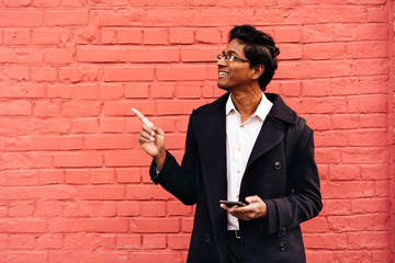 A man in a dark blue jacket is standing in city by red pink wall with a phone in his hands looking for navigation. Solo overseas traveler, sightseeing. Taxi booking by mobile app