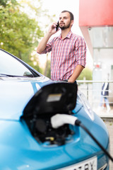 Handsome young guy with a beard talking on the phone near his electric car.