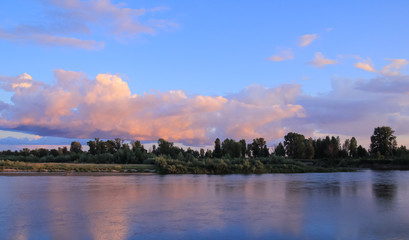 Sunset on the river with the bank covered by small forest and blue-pink sky