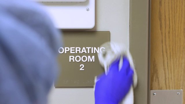 Female janitor cleaning brail sign for operating room 2 in hospital doctors office and sanitizing the sign 
