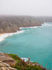 Wide aerial view of the rocky granite cliffs along the foggy coasts of Porthcurno Beach. Vertical orientation. Cornwall, United Kingdom. Travel and nature. - 310677353