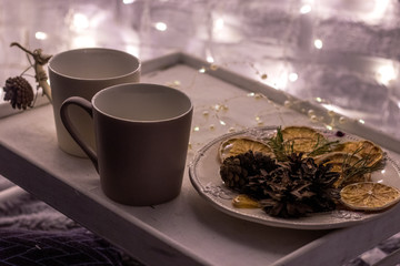 two white mugs on a wooden tray stand on a bed with a fluffy blanket with a garland in the background