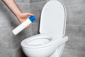 Cropped view of man holding air freshener above toilet bowl in bathroom with grey tile