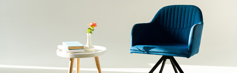 Panoramic shot of blue armchair by coffee table with flower in vase and books on grey background