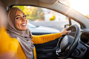 Young smiling Muslim gril making selfie portrait sitting in the car. Beautiful young Muslim woman in car, taking selfie. Road trip selfie. Young woman using her smartphone and making selfie