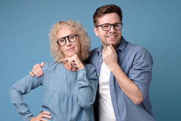 Two mature man and woman in glasses and casual clothes looking confident at camera standing on blue wall. Positive facial human emotion. Studio shot
