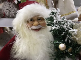 Toy Santa Claus in glasses with a white fluffy beard and a hat in a red suit on the background of a Christmas tree and other toys in the store. Photo with mobile under shop lighting. 