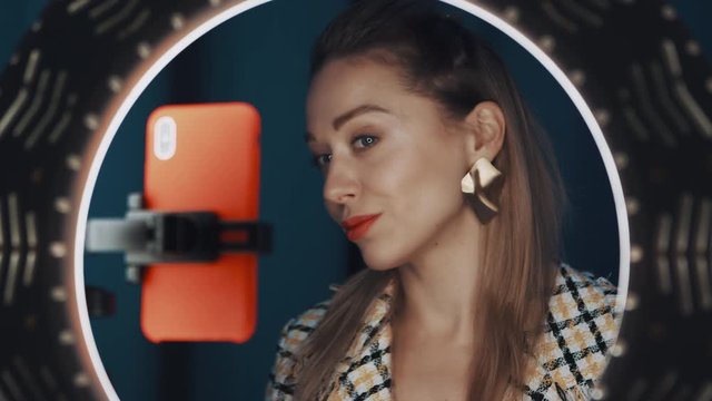 Young nice-looking woman beauty blogger sits in front of ring light and orange smartphone, touches her lips with finger, waving good-bye to camera, recording tutorial video. Shot on 4K RED camera.