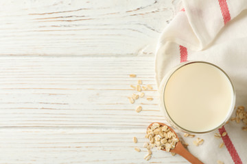 Glass of oat milk, spoon with oatmeal seeds and napkin on white, wooden background, top view. Space for text