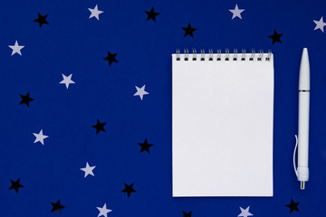 white notebook and pen for entries on classic blue background with stars. top view, colour trend 2020