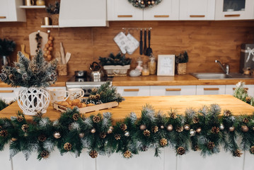 View over beautiful white kitchen with Christmas decorations all over cupboards and kitchen board. There is Christmas wreath on the cupboard. Natural fir tree branches with pine cones.