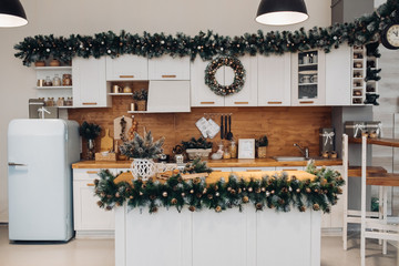 View over beautiful white kitchen with Christmas decorations all over cupboards and kitchen board. There is Christmas wreath on the cupboard. Natural fir tree branches with pine cones.