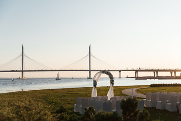 arch for the wedding ceremony and chairs for guests stand on the background of water and a bridge