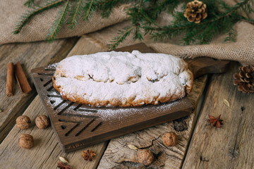Homemade traditional dessert stollen with powdered sugar on top - stands on rustic wooden board with pine nuts, anise, sackcloth, fir tree branches decoration.
