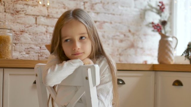 Little sad girl with blue eyes and long blond hair wearing white dress and sweater sits on a chair in a loft style room