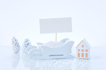 White holiday scene with a white paper card on sledges with white cones and a paper house. Christmas scene arrangement.