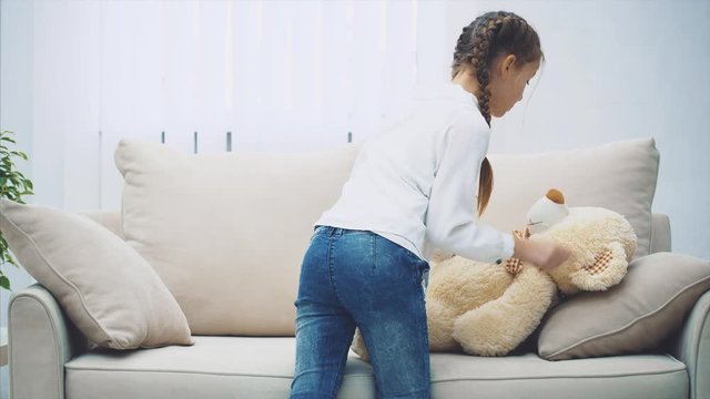Cute little girl putting white teddy on the sofa, petting it, going away.