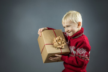 Cute cheerful child in a Christmas sweater holds a big gift wrapped in kraft paper. The boy enjoys a lot of fun.