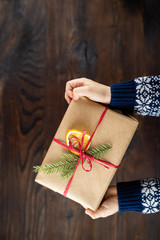 Childers hands hold a present box. Gift wrapped in kraft paper and decorated with a spruce branch and dry orange. Christmas presents on a wooden table with a copy space.