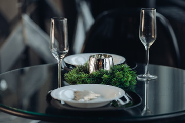 View over beautifully decorated round table with natural fir branch, candle, two flutes, plates against classic sofa in modern apartment.