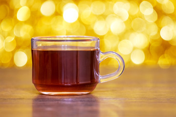 Glass transparent cup with tea on a wooden table on a bright golden background with a bright bokeh_