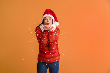 Young woman in winter clothes blowing snow on color background