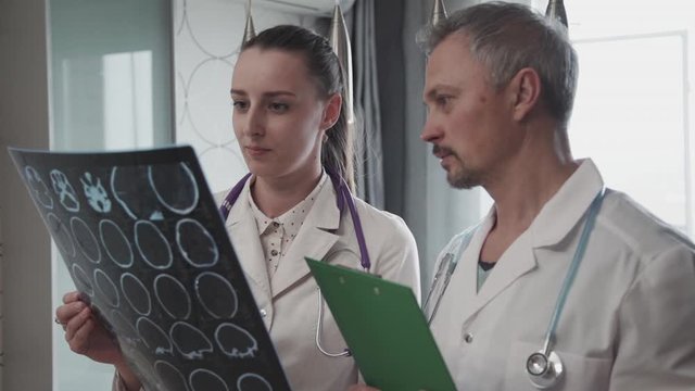 Serious mature doctor and young adult nurse holding x ray picture in hands, making conversation about treatment and working at office in clinical laboratory, wearing special uniform