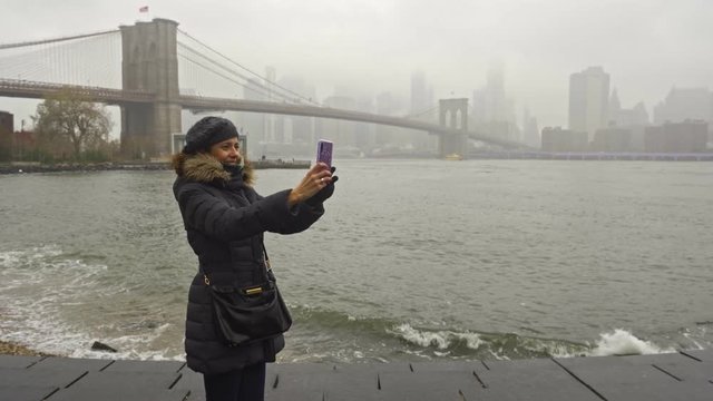 Beautiful curly brunette woman taking selfie self-portrait in front of the Brooklyn Bridge over East River, while sightseeing new york during winter season