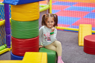 Fototapeta na wymiar Little cute caucasian girl with funny ponytail playing with soft building blocks at indoor playground, sitting and looking directly at camera, spending time happily, wearing blue casual sweater.