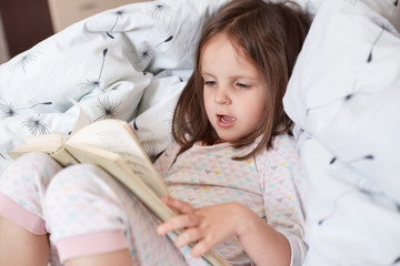 Close up portrait of little girl reading story or fairy tale for her teddy bears while laying in bed and wearing pajama, charming kid speling words aloud, looks concentrated, being in good mood.