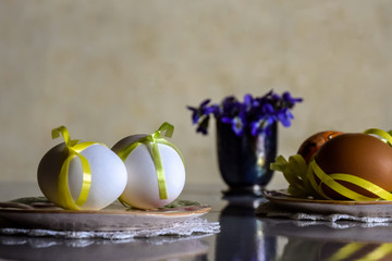 Easter setting: two ceramic plates with white and brown eggs, decorated with ribbons, and bouquet of blue forest flowers in small vase are mirrored on the table surface.