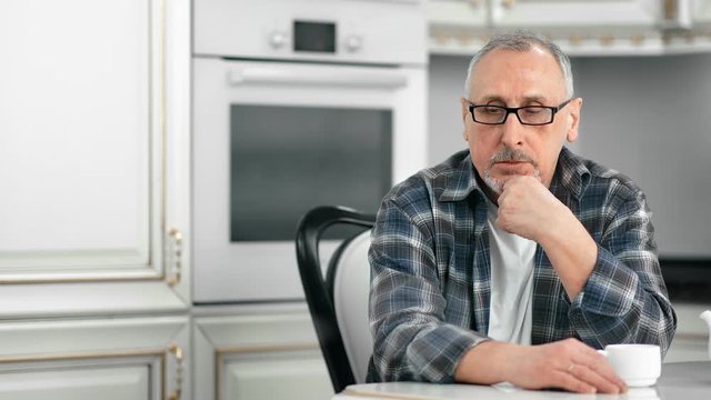 Portrait of pensive gray-haired casual elderly man sitting at white kitchen during coffee break