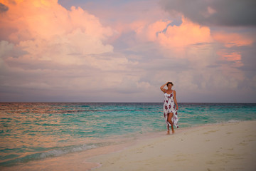 Young lonely woman in colorful dress walking on the seashore, looking at turquoise ocean water on luxury tropical island resort. Beautiful sunset evening.