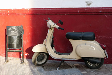 Old cream colored vespa / motor scooter next to rubbish bin in front of red wall