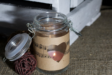 Glass jar wrapped by paper with notes. Heart cut out in paper. It is on a linen cloth. A candle is burning in the bank. Near rattan balls and a wooden box of boards.