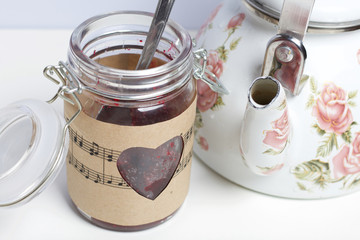 Glass jar of jam wrapped in paper with notes. Heart cut out in paper. Near a white kettle with roses.