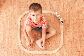 Cute little boy sitting on floor, around him is wooden railway with a train. Child looking at camera. Top view.