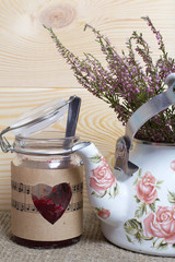 Glass jar of jam wrapped in paper with notes. Heart cut out in paper. Near the kettle with a bunch of heather.