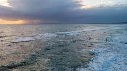 An aerial view of a choppy sea with crashing waves and groynes (breakwater) under a stormy cloudy grey sky with ray of light from the sun behind it.