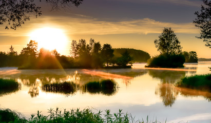 Fototapeta premium Sunrise, a large white sun over the mirror surface of the river, small green islands with trees, the rays of the sun, morning in the summer, Plavinas Latvia.