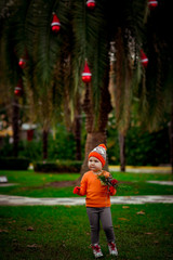 a small, beautiful girl in bright clothes,and in an orange hat, dancing near a Christmas tree, with a Christmas bouquet in her hand