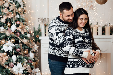 Attractive cheerful man and woman near christmas tree hugging each other. New Year's eve. Young happy  pregnant couple. Family, christmas, winter, happiness and people concept