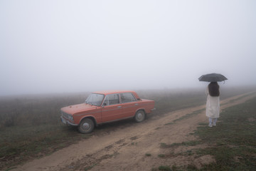 Red retro car stands near the dirt road in fog, young girl in white coat stands under umbrella