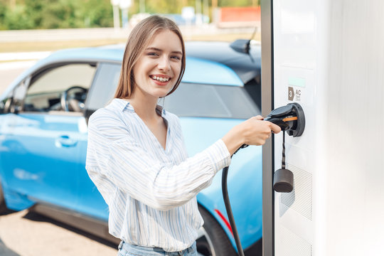 Transportation. Young woman on electric car having stop at charging station standing with charger smiling cheerful